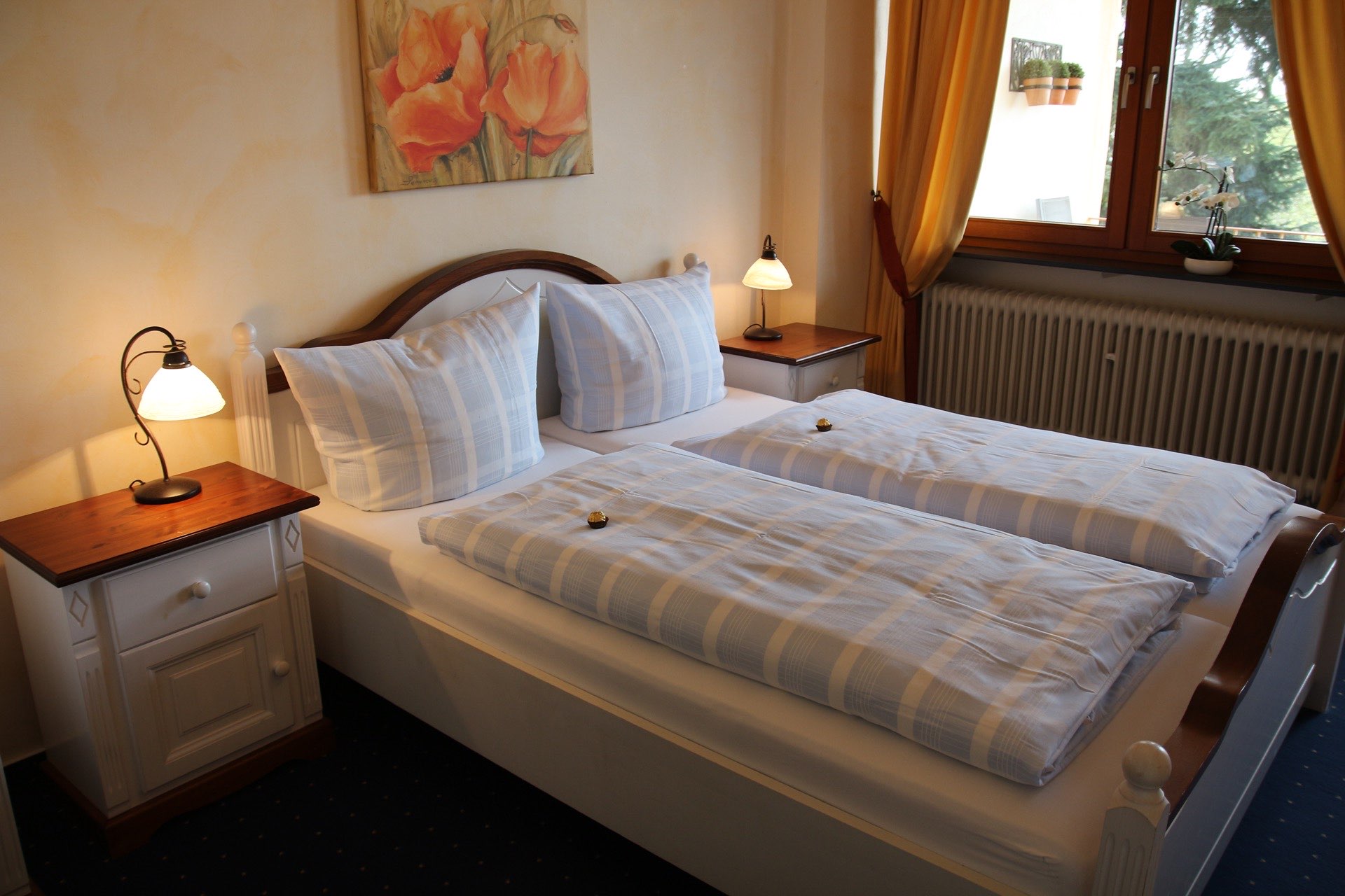 Helles Doppelzimmer ‚Lucca‘ in der Pension Rehwinkel in Nalbach, mit weißen Möbeln, warmen Gelbtönen, künstlerischem Blumenbild und einem großen Fenster mit Vorhängen.